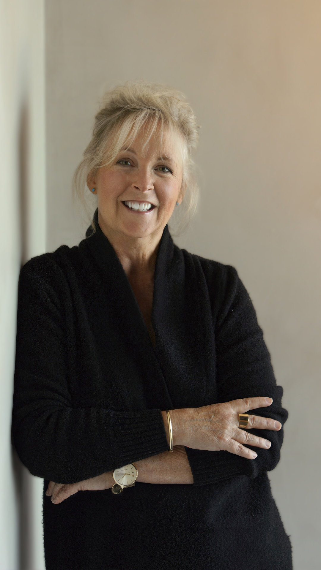Jami McNees, a woman with blonde hair, wearing a black sweater and gold jewelry, leaning on a wall with folded arms and a warm smile.